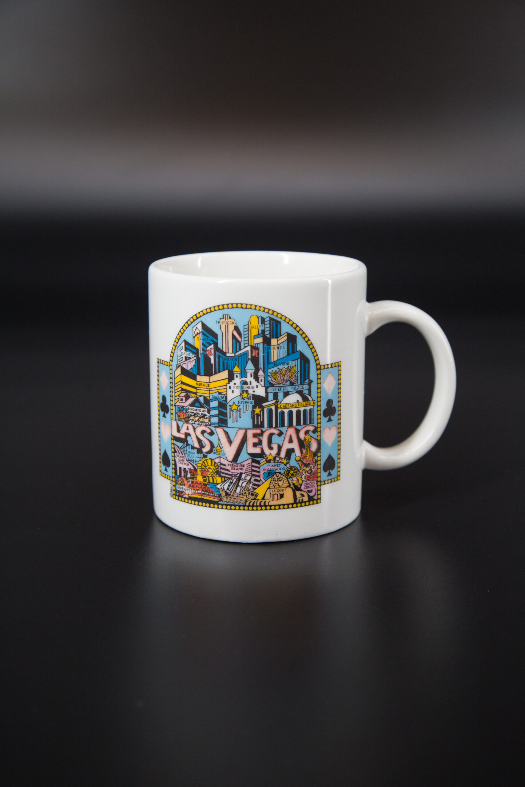 Tazza in ceramica bianca con stampa colorata dello skyline di Las Vegas e simboli delle carte da gioco, su sfondo nero.