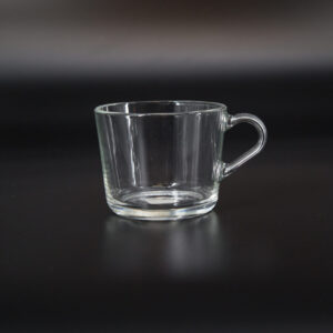 Tazza in vetro trasparente con manico, fotografata su sfondo scuro. L'oggetto è vuoto e riflette la luce.