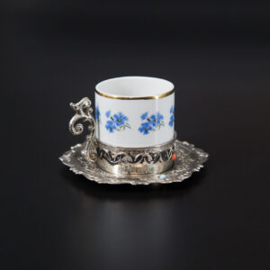 Tazzina da caffè in porcellana bianca con fiori blu e bordo oro, inserita in un supporto e piattino in metallo argentato ornato.