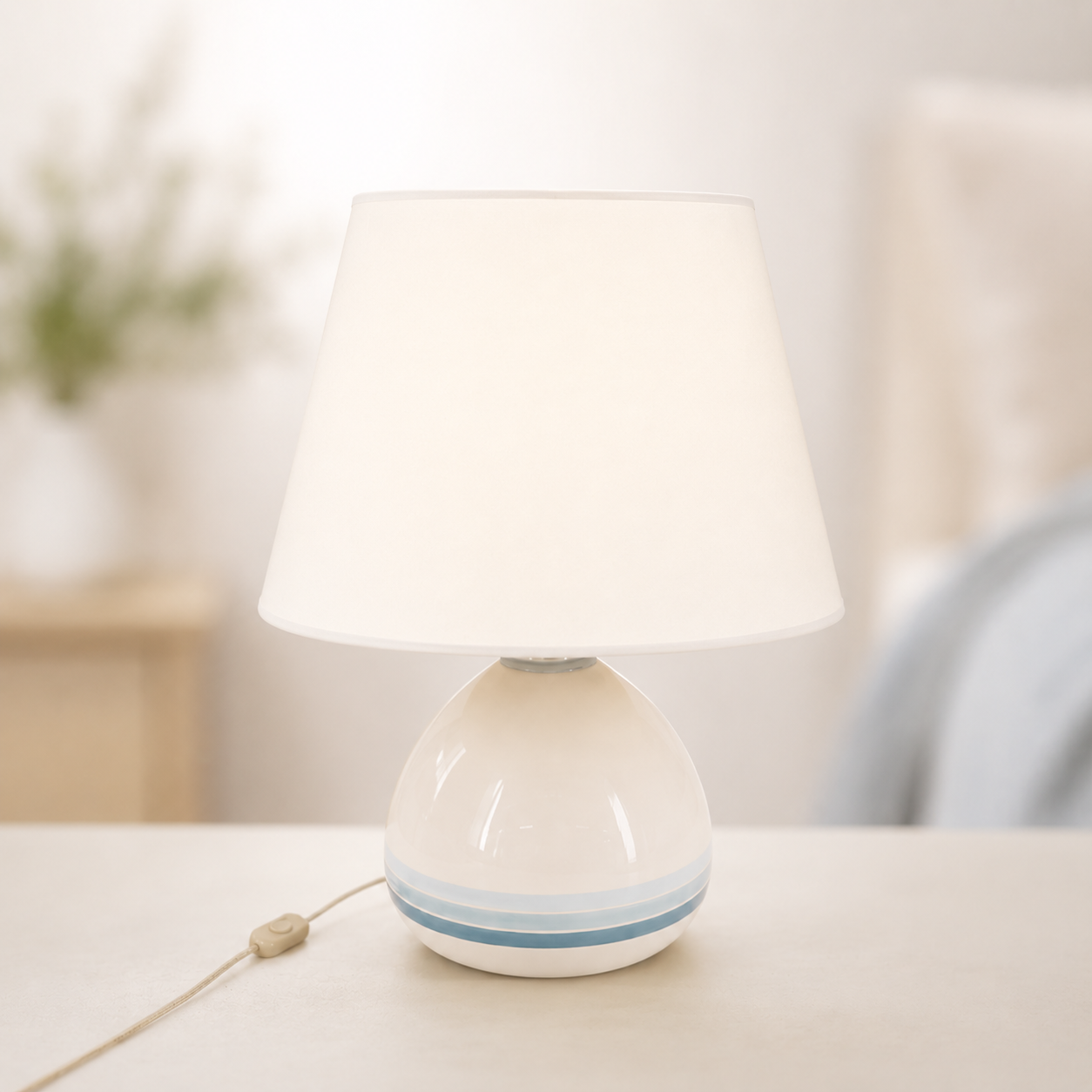 Lampada da tavolo con base in ceramica bianca a righe blu e paralume conico bianco crema, su un tavolo chiaro.