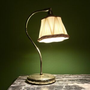 Lampada da tavolo vintage con base e stelo in ottone lucido e paralume plissettato color crema con bordo decorato, accesa su un tavolo di marmo scuro.