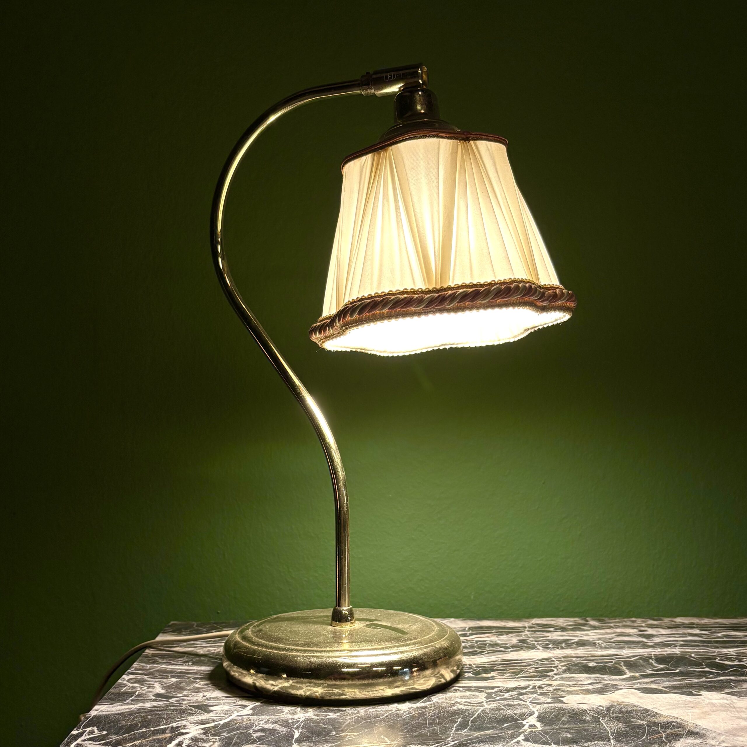 Lampada da tavolo vintage con base e stelo in ottone lucido e paralume plissettato color crema con bordo decorato, accesa su un tavolo di marmo scuro.