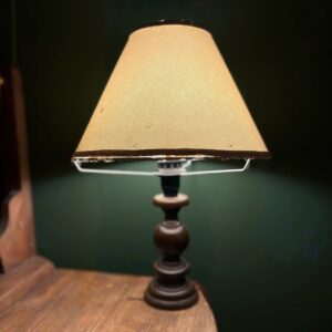 Lampada da tavolo vintage accesa con base in legno tornito scuro e paralume conico in tessuto beige, su superficie in legno.