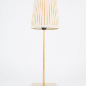 Lampada da tavolo moderna con base e fusto in ottone spazzolato e paralume conico plissettato color crema su sfondo bianco.