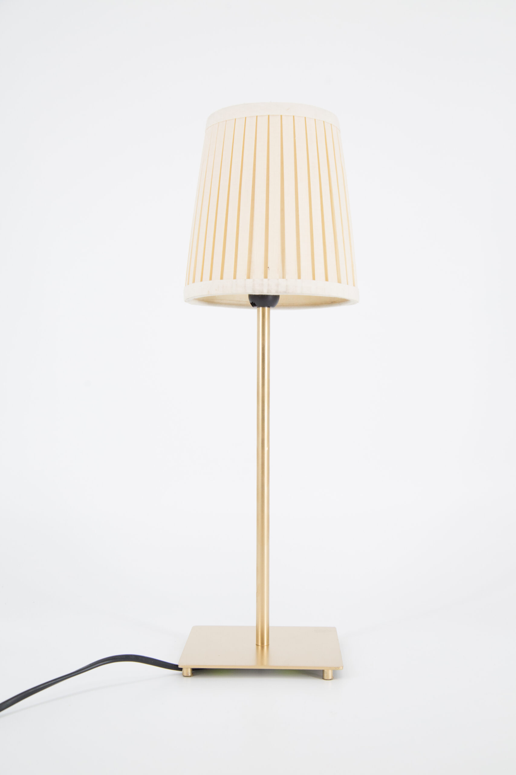 Lampada da tavolo moderna con base e fusto in ottone spazzolato e paralume conico plissettato color crema su sfondo bianco.