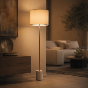 Lampada da terra moderna con paralume in tessuto beige, stelo sottile color ottone e base cilindrica in marmo bianco, accesa in un ambiente interno.