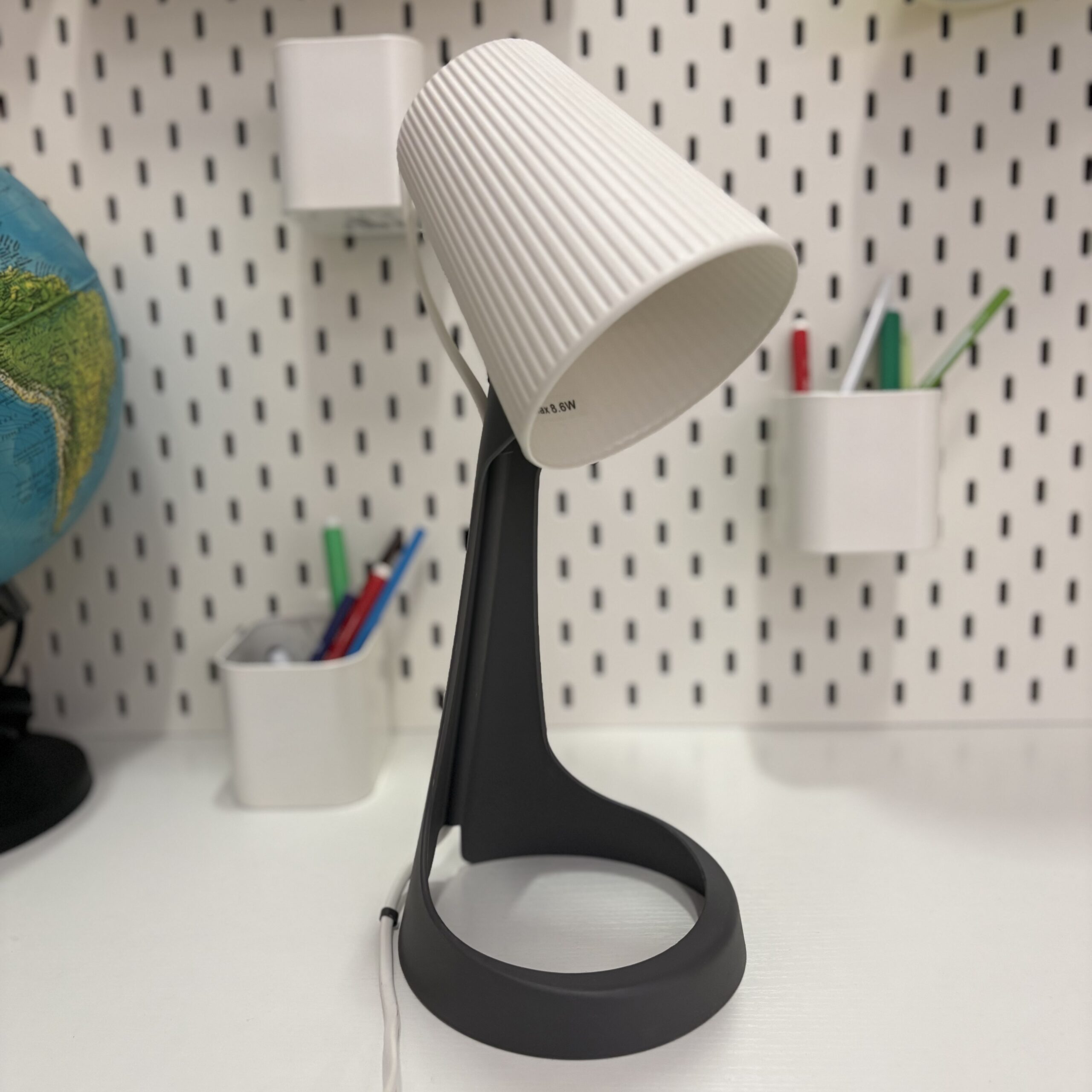 Lampada da tavolo moderna con base grigio scuro e paralume bianco a coste, su scrivania bianca con pegboard.