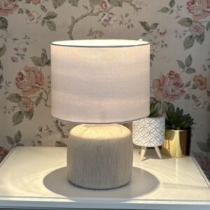 Lampada da tavolo con base cilindrica beige testurizzata e paralume in tessuto chiaro acceso, su un comodino bianco con sfondo floreale.