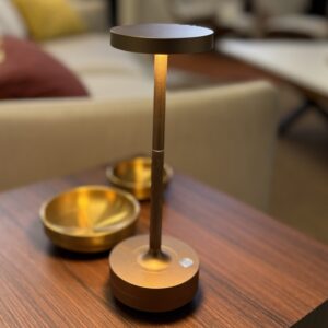 Lampada da tavolo portatile LED color bronzo con design moderno, accesa su un tavolino di legno scuro, con ciotole dorate sullo sfondo sfocato.