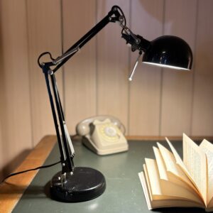 Lampada da tavolo articolata nera accesa su una scrivania verde, con un libro aperto e un telefono a disco beige sullo sfondo.