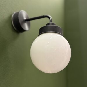 Applique da parete con braccio curvo nero opaco e diffusore sferico in vetro opalino bianco, montata su parete verde.