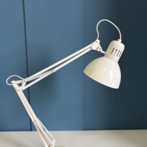 Lampada da tavolo articolata bianca con braccio a molle e testa a campana, su sfondo blu.