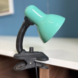 Lampada da tavolo a pinza con paralume verde acqua e braccio flessibile nero, fissata su un bordo bianco.