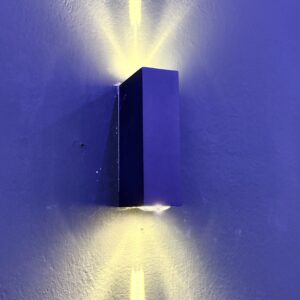 Applique da parete nera rettangolare montata su una parete blu, che emette fasci di luce gialla verso l'alto e verso il basso.
