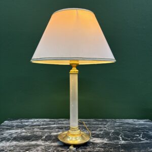 Lampada da tavolo classica con base e dettagli dorati, fusto scanalato trasparente e paralume conico bianco, su un tavolo di marmo.
