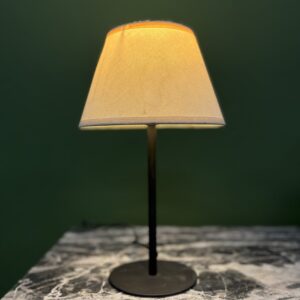 Lampada da tavolo accesa con paralume conico in tessuto beige e base scura, su superficie marmorizzata.
