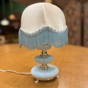 Lampada da tavolo vintage con base azzurra lucida, dettagli dorati e paralume in tessuto crema con frange azzurre.