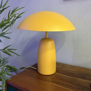 Lampada da tavolo gialla opaca con paralume a cupola e base cilindrica, posizionata su un mobile in legno.