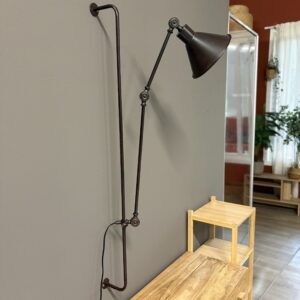 Lampada da parete industriale con braccio articolato e paralume a cono, finitura metallica anticata, montata su parete grigia.