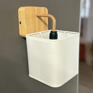 Applique da parete moderna con base in legno chiaro e paralume quadrato in tessuto bianco, fissata su una parete grigia.