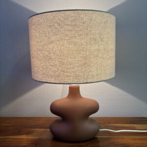 Lampada da tavolo accesa con base scultorea in terracotta marrone e paralume cilindrico in tessuto beige su tavolo di legno.
