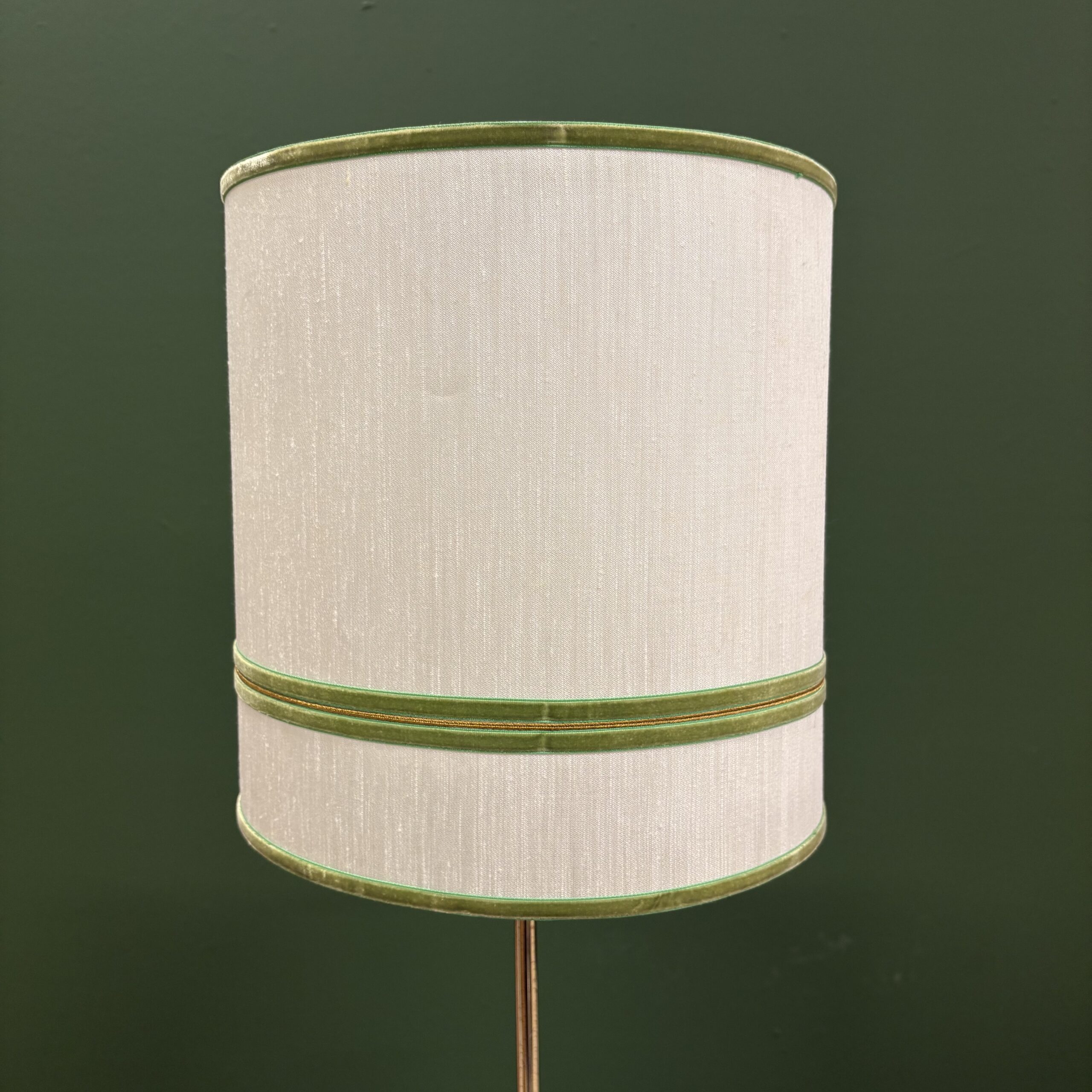 Paralume cilindrico in tessuto color crema con bordi e fascia centrale in velluto verde e cordino dorato, su sfondo verde scuro.