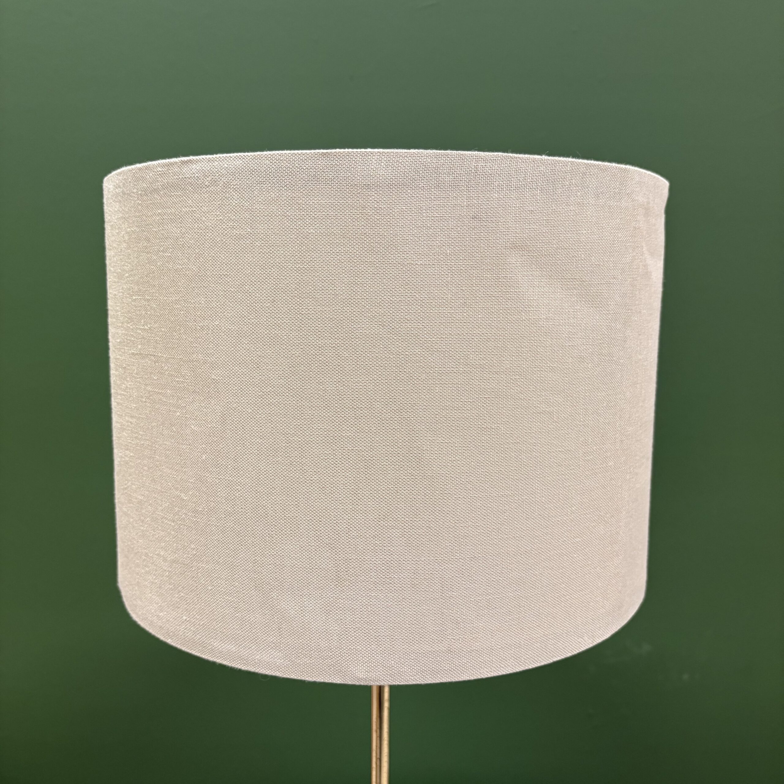 Paralume cilindrico in tessuto beige chiaro con trama visibile, montato su un sottile stelo dorato, su sfondo verde scuro.