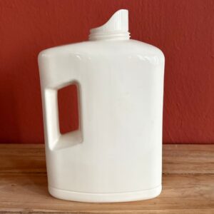 Flacone generico in plastica bianca con manico, senza tappo, su superficie in legno con sfondo rosso-arancio.