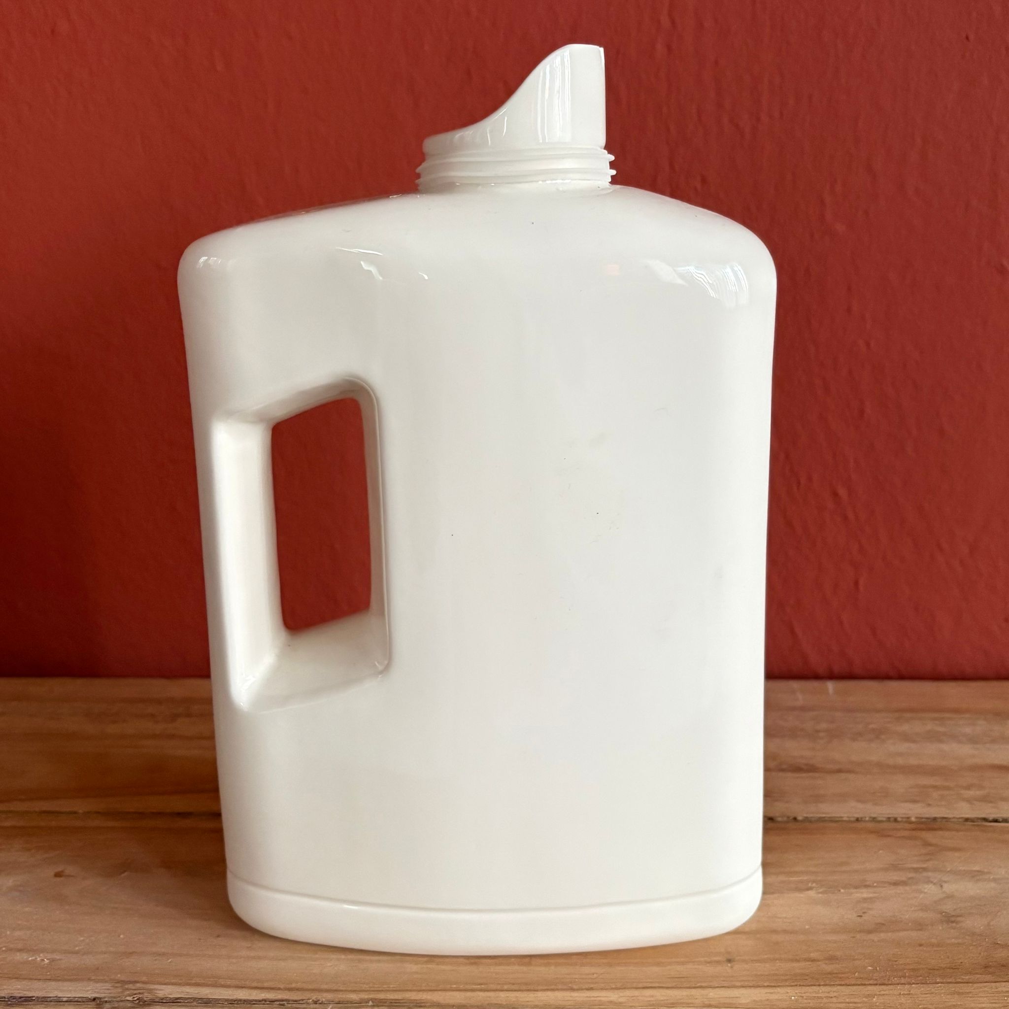 Flacone generico in plastica bianca con manico, senza tappo, su superficie in legno con sfondo rosso-arancio.