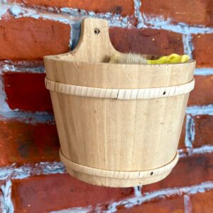 Secchio in legno chiaro con fasce decorative e manico, contenente una spazzola e un panno giallo, appeso a una parete di mattoni rossi.