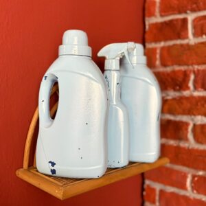 Tre flaconi di plastica chiari, dall'aspetto invecchiato e scrostato, su una mensola di vimini contro una parete rossa e mattoni.