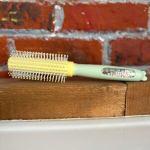 Spazzola rotonda con corpo giallo e setole bianche, manico verde chiaro con inserto trasparente contenente perline colorate, su legno.