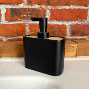 Dispenser di sapone nero opaco con top in legno chiaro e pompa nera, posizionato su una superficie bianca davanti a un muro di mattoni rossi.