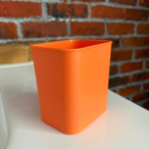 Un contenitore quadrato di colore arancione brillante, realizzato in plastica, posizionato su una superficie bianca con uno sfondo di mattoni rossi.