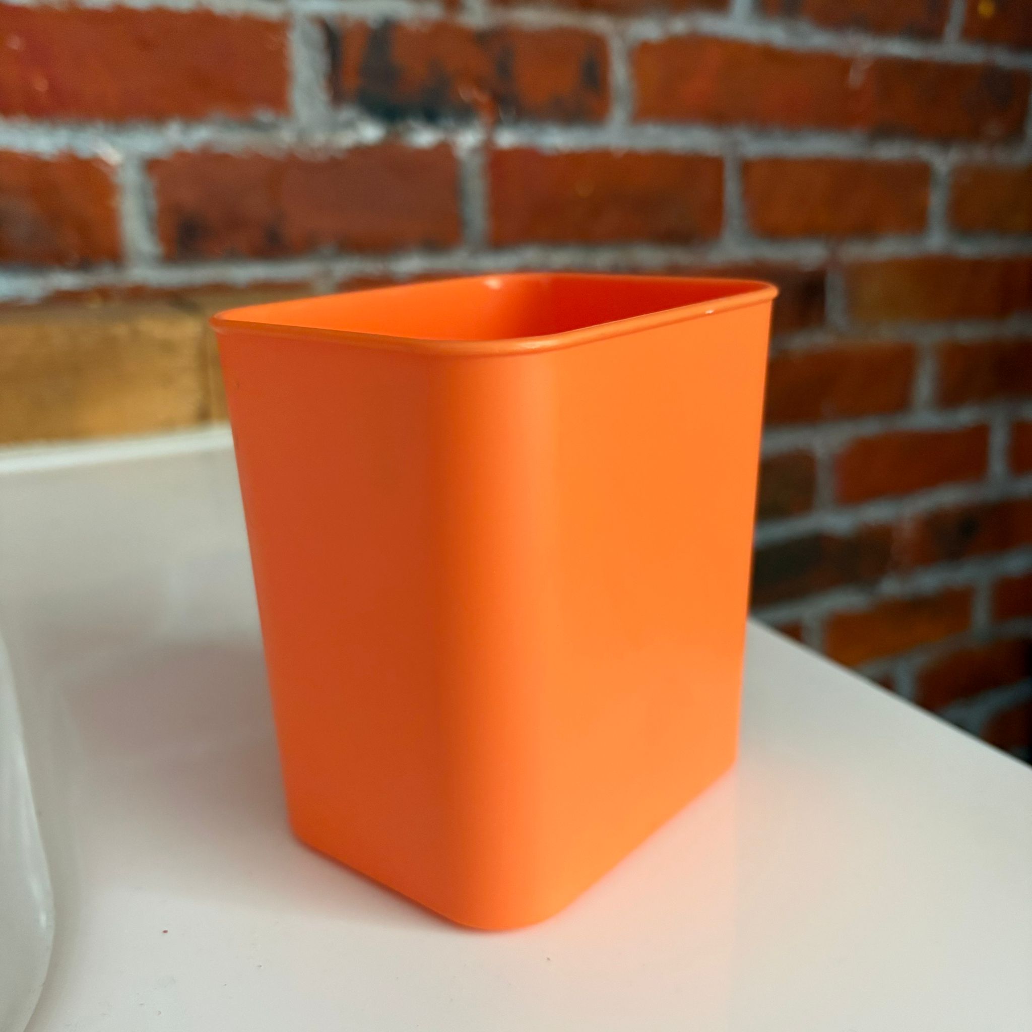Un contenitore quadrato di colore arancione brillante, realizzato in plastica, posizionato su una superficie bianca con uno sfondo di mattoni rossi.