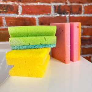 Un set di cinque spugne da cucina in schiuma, di colori verde, giallo, rosa e arancione, appoggiate su una superficie bianca con uno sfondo di mattoni rossi.