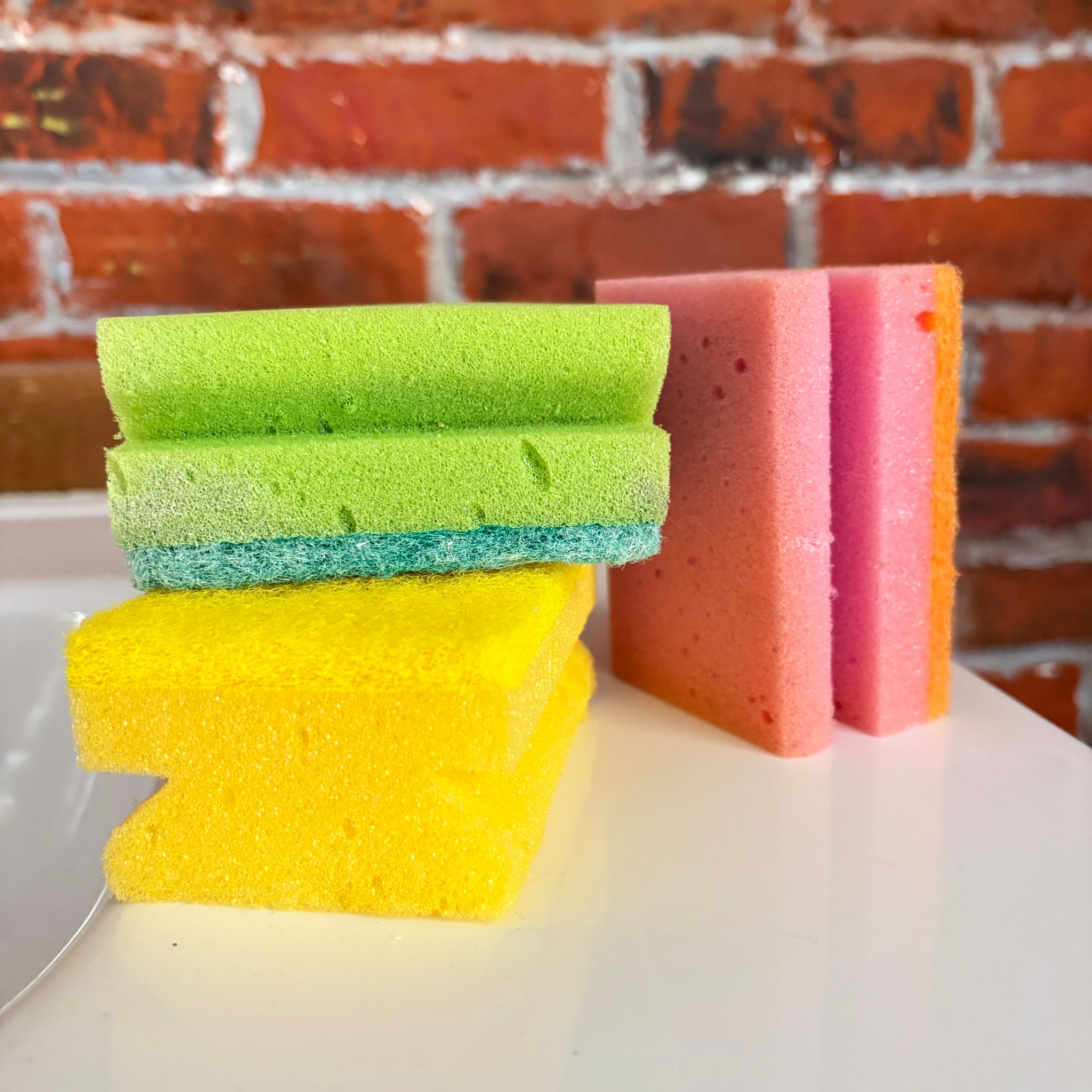 Un set di cinque spugne da cucina in schiuma, di colori verde, giallo, rosa e arancione, appoggiate su una superficie bianca con uno sfondo di mattoni rossi.