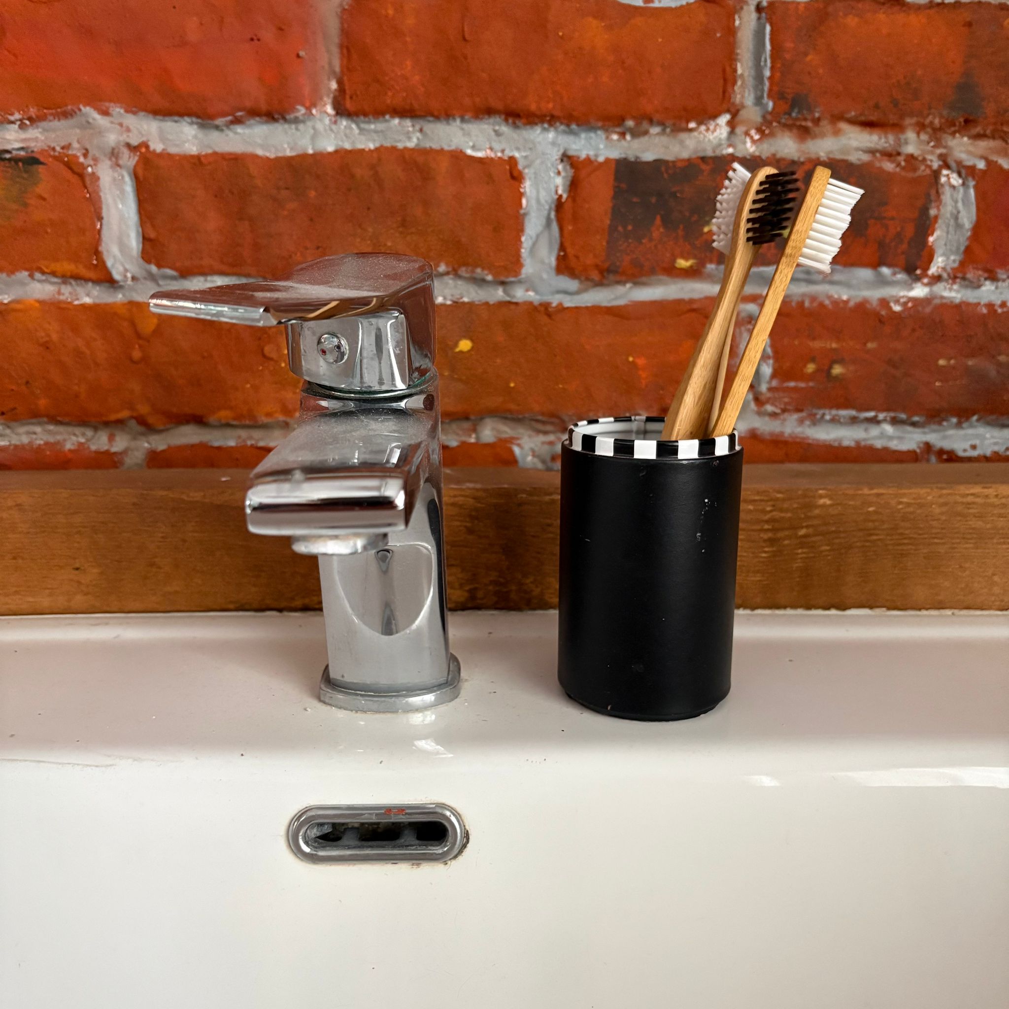 Due spazzolini in bambù in un porta spazzolini nero a righe, posizionati su un lavabo bianco con rubinetto cromato e parete in mattoni rossi.