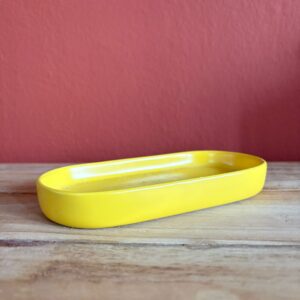 Un vassoio ovale di colore giallo brillante e lucido, posizionato su una superficie di legno chiaro con uno sfondo rosso-arancio.