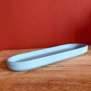 Vassoio ovale allungato di colore azzurro pastello con finitura opaca, posizionato su una superficie di legno chiaro davanti a una parete rossa.
