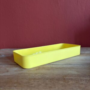 Vassoio rettangolare giallo brillante in plastica, posizionato su una superficie di legno chiara con sfondo rosso bordeaux.