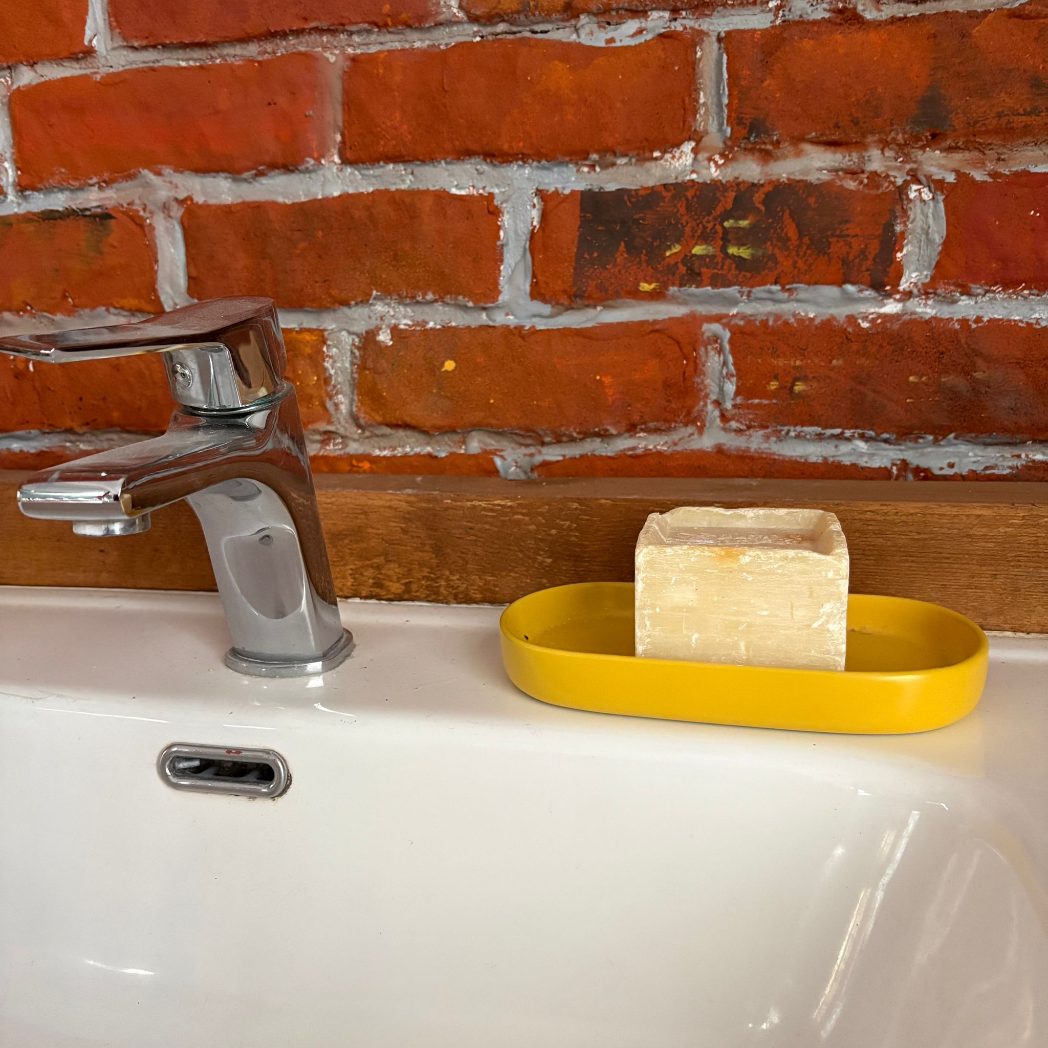 Rubinetto monocomando cromato su lavabo bianco con porta sapone giallo e saponetta, su sfondo di parete in mattoni.