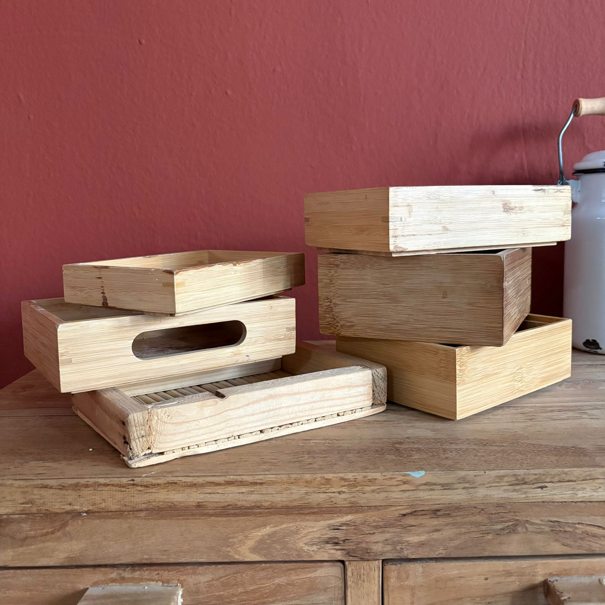 Sei contenitori e vassoi in legno chiaro di diverse forme e dimensioni, impilati su un mobile in legno contro una parete rossa.