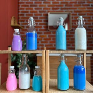 Otto bottiglie di vetro di varie forme e dimensioni, riempite con liquidi opachi colorati (rosa, viola, azzurro, blu, bianco), disposte su uno scaffale di legno.
