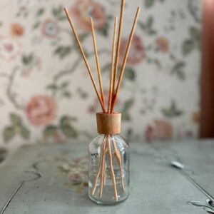 Diffusore a bastoncini vuoto con bottiglia in vetro trasparente, tappo in legno chiaro e bastoncini di rattan su tavolo.