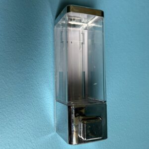 Dispenser da parete rettangolare con serbatoio trasparente e base cromata lucida, montato su una superficie blu.