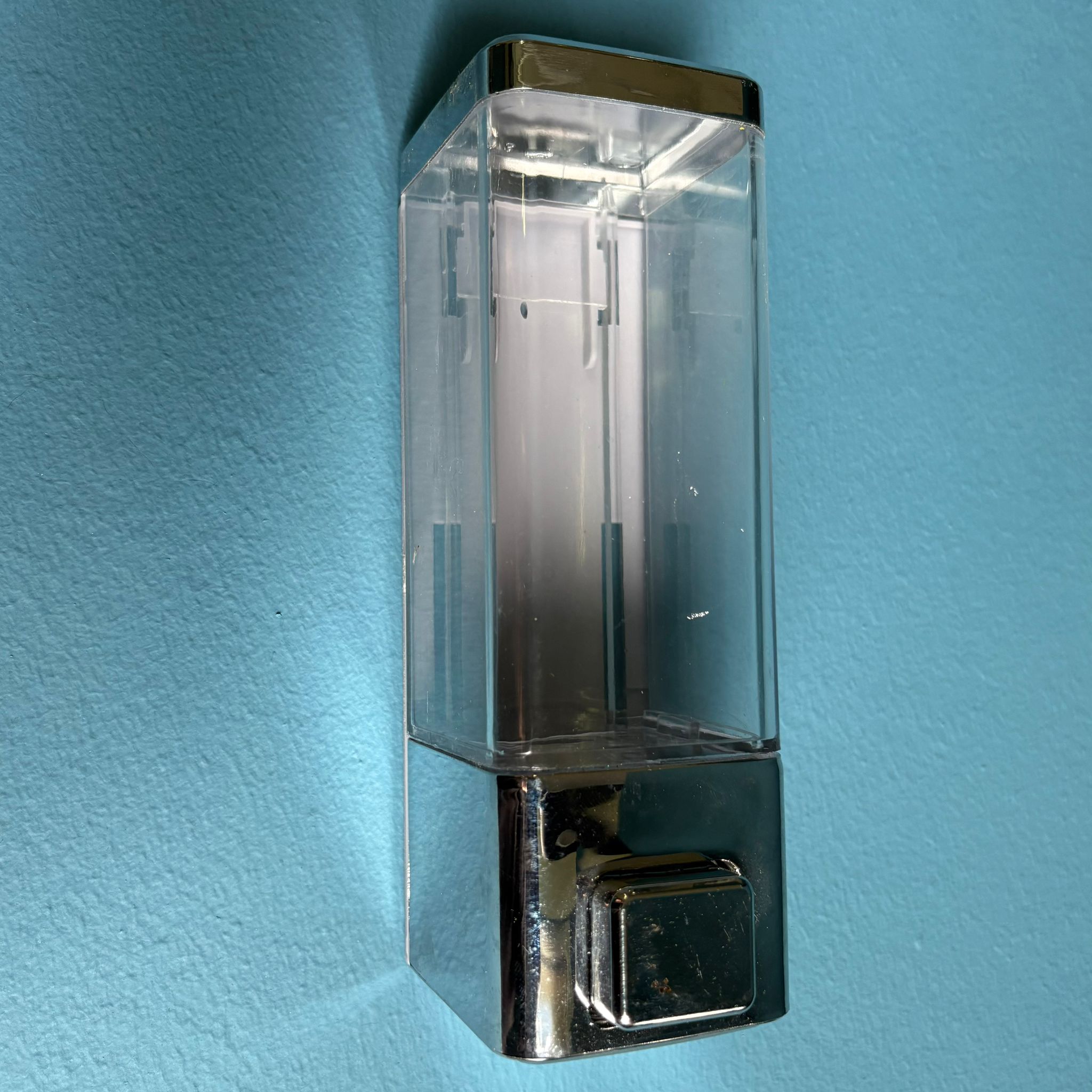 Dispenser da parete rettangolare con serbatoio trasparente e base cromata lucida, montato su una superficie blu.