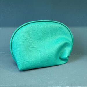 Pochette cosmetica semicircolare di colore teal in similpelle testurizzata, posizionata su una superficie grigio-blu.