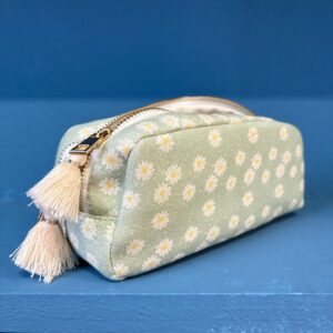 Astuccio rettangolare in tessuto verde menta con fantasia a margherite bianche, cerniera dorata e due nappe color crema, su sfondo blu.