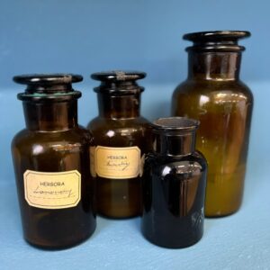 Quattro flaconi da farmacia antichi di vetro ambrato e nero, con tappi a smeriglio e etichette "HERBORA", su sfondo blu.